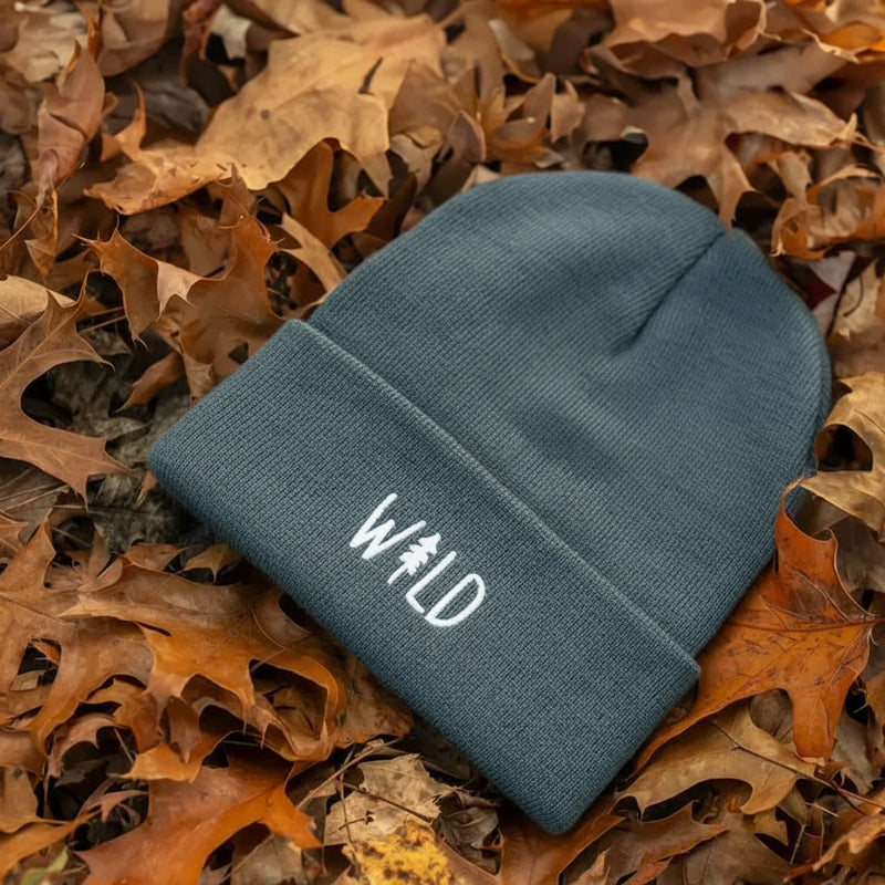 Gray beanie with white WILD text and a tree graphic, resting on brown autumn leaves