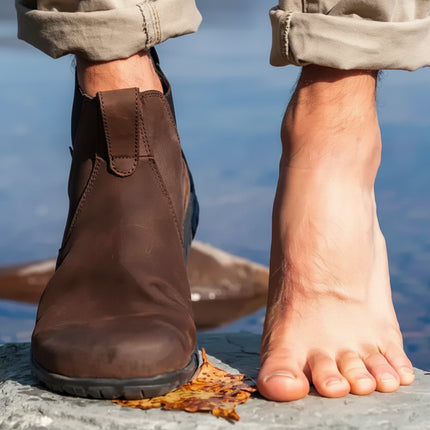 A person stands with one foot in a brown boot and the other bare, on a rock by water