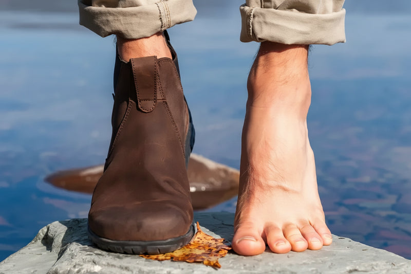 A person stands with one foot in a brown boot and the other bare, on a rock by water