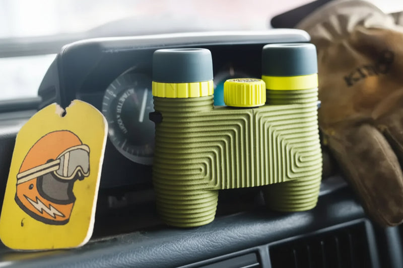 Green binoculars with yellow accents on a car dashboard next to a helmet air freshener and a glove