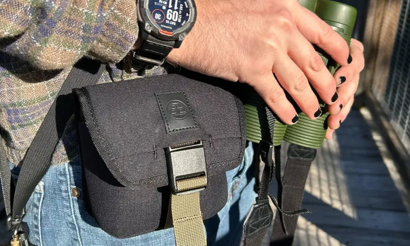 Person wearing a black shoulder bag, a watch, and holding green binoculars