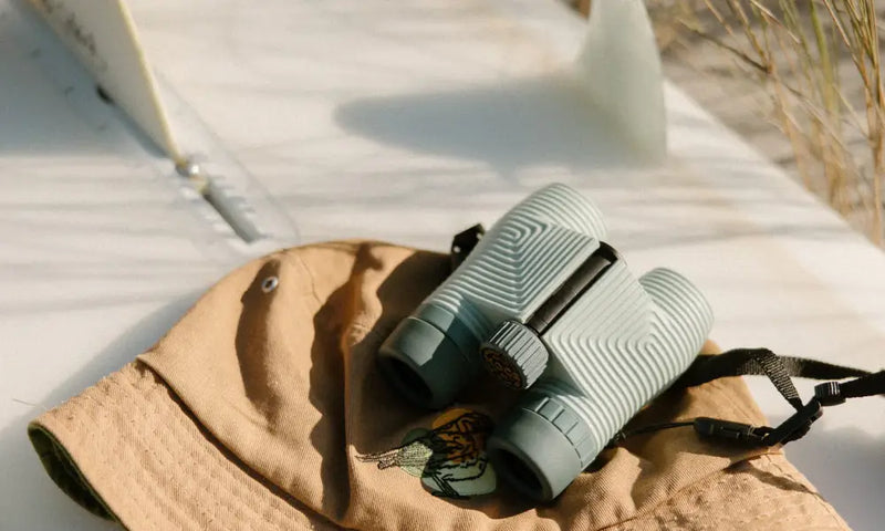 Light blue binoculars with a black strap resting on a tan bucket hat with an embroidered bird