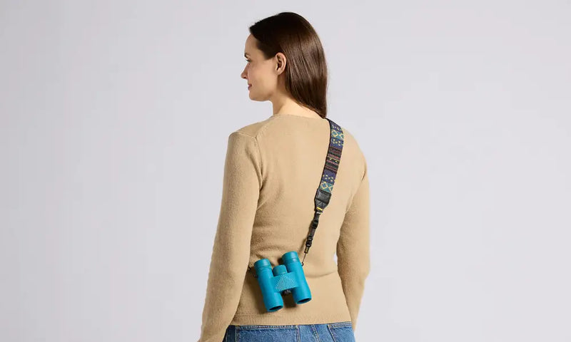 Woman wearing a beige sweater and blue jeans with turquoise binoculars hanging from a patterned strap