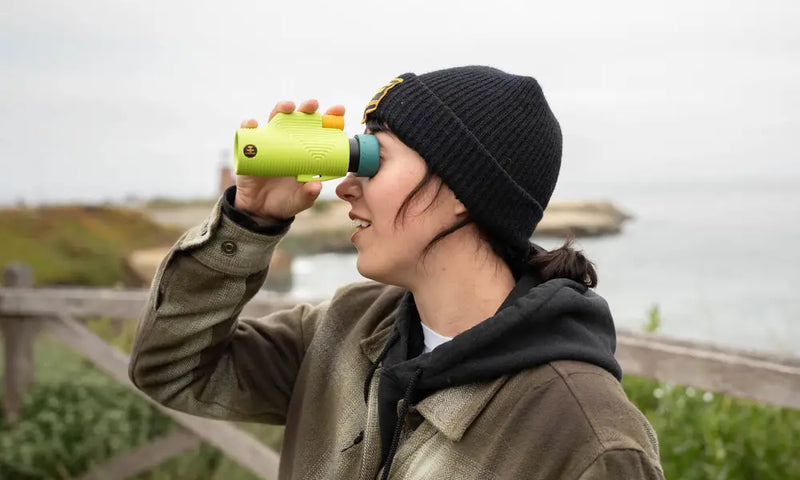 Person in a black beanie and brown jacket looking through green binoculars at the ocean