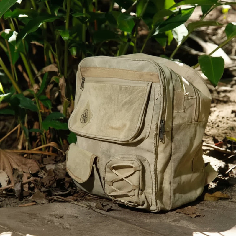 Light brown canvas backpack with multiple pockets and zippers, resting on a wooden surface with green foliage
