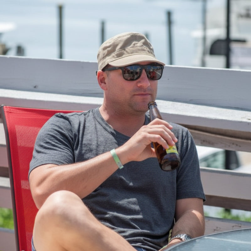 Man in a grey t-shirt, sunglasses, and a tan hat drinking from a brown bottle