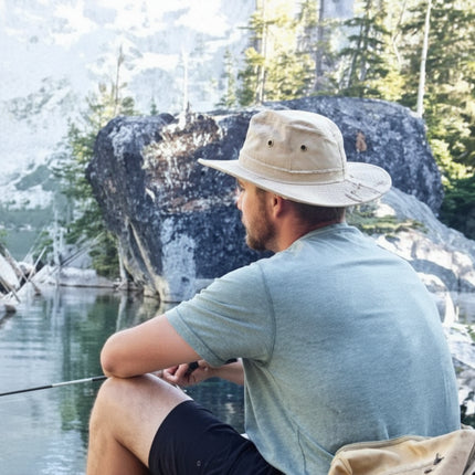Man in a wide-brimmed hat fishing by a lake with mountains and trees in the background