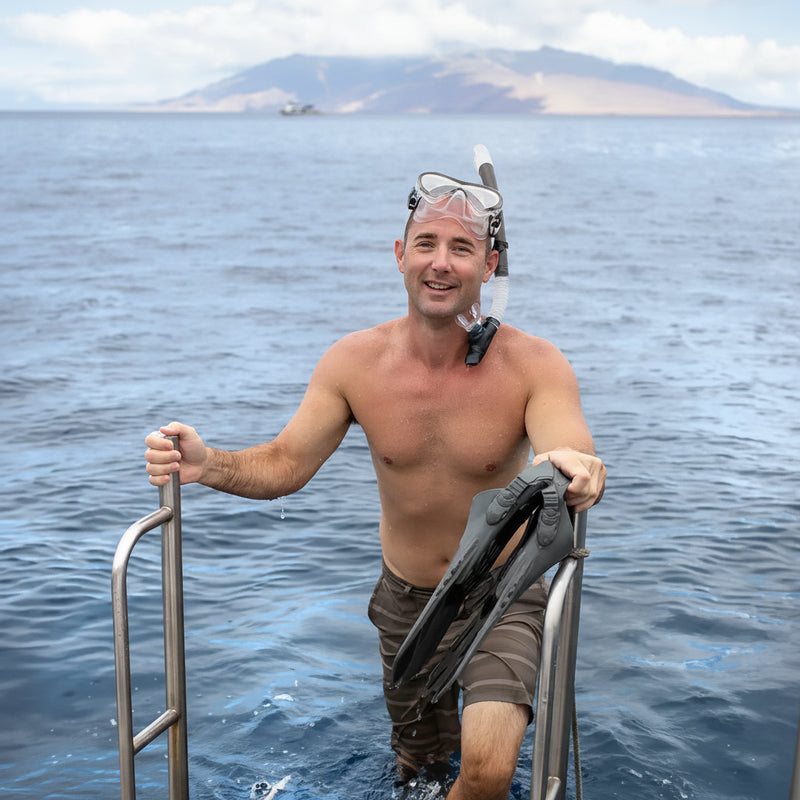 Man in snorkel gear and swim trunks climbing out of the ocean onto a boat with seavenger snorkel and fins.