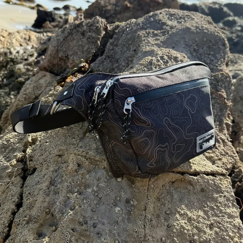 Black fanny pack with a topographic map pattern and silver zippers, resting on a rock