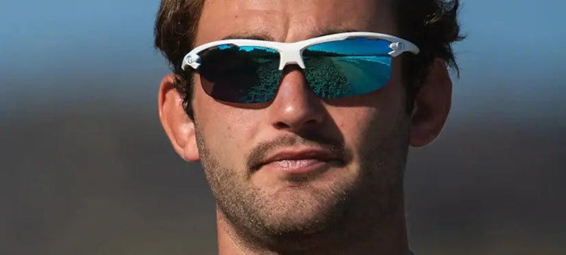 Man wearing white sunglasses with blue reflective lenses, showing a beach scene reflection