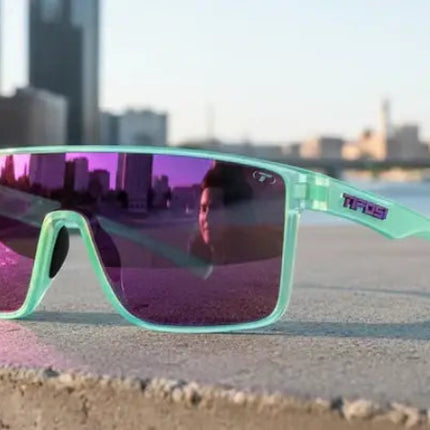 Mint green sunglasses with purple lenses on a concrete ledge, city skyline reflected in lenses