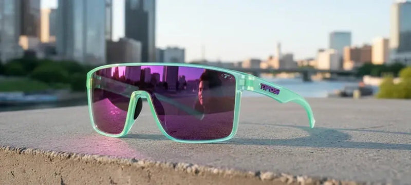 Mint green sunglasses with purple lenses on a concrete ledge, city skyline reflected in lenses
