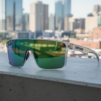 Tifosi sunglasses with clear frames and green mirrored lenses on a concrete ledge with a city in the background