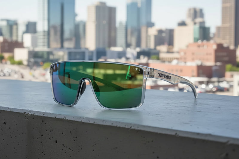 Tifosi sunglasses with clear frames and green mirrored lenses on a concrete ledge with a city in the background
