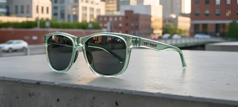 Light green sunglasses with dark lenses on a concrete ledge, city buildings and a car in the background