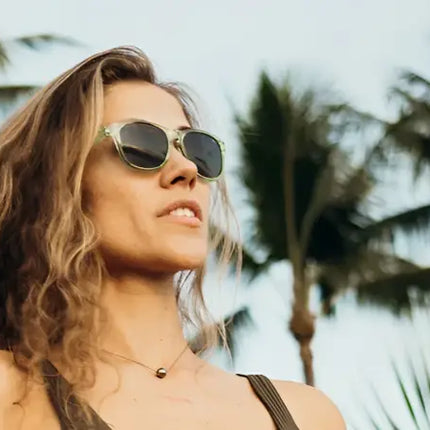 Woman with curly hair wearing sunglasses and a necklace, with palm trees in the background