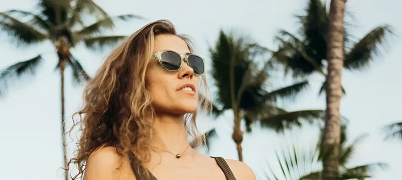 Woman with curly hair wearing sunglasses and a necklace, with palm trees in the background
