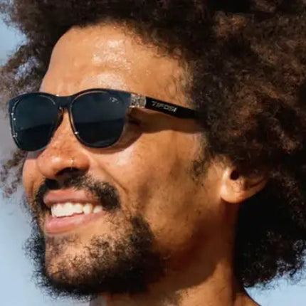 Man with dark curly hair and beard wearing black sunglasses with a subtle logo on the side