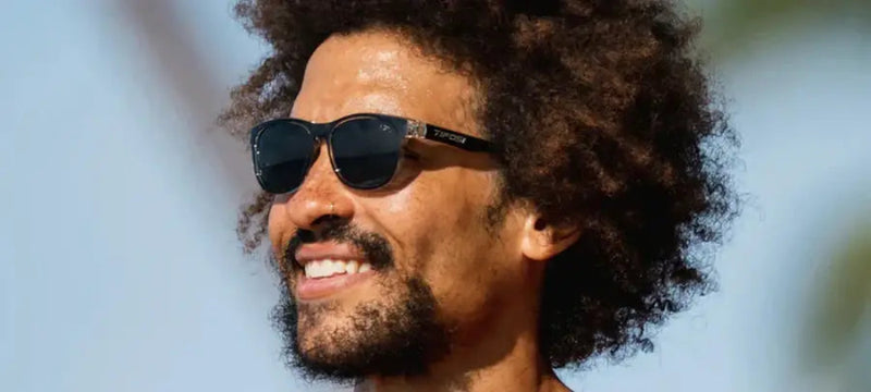 Man with dark curly hair and beard wearing black sunglasses with a subtle logo on the side
