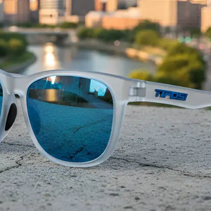 Frosted white sunglasses with blue mirrored lenses on a concrete ledge overlooking a city and river