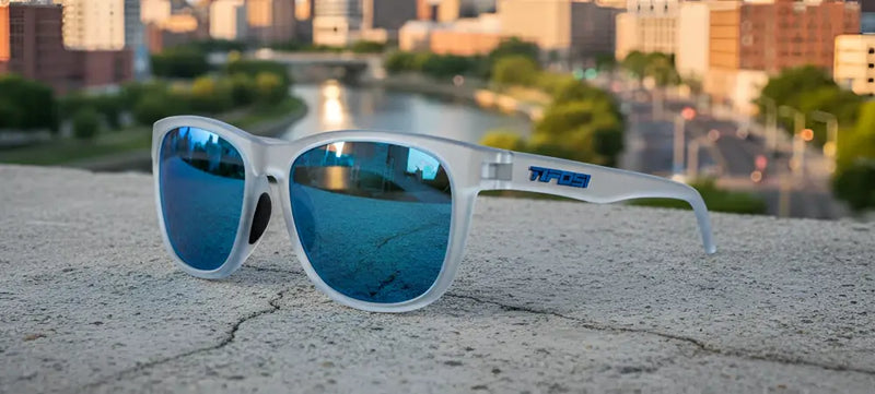 Frosted white sunglasses with blue mirrored lenses on a concrete ledge overlooking a city and river