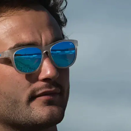 Man wearing clear frame sunglasses with blue mirrored lenses, reflecting a beach scene
