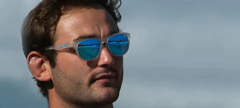 Man wearing clear frame sunglasses with blue mirrored lenses, reflecting a beach scene