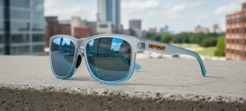 Light blue sunglasses with reflective blue lenses on a concrete ledge, city buildings in background