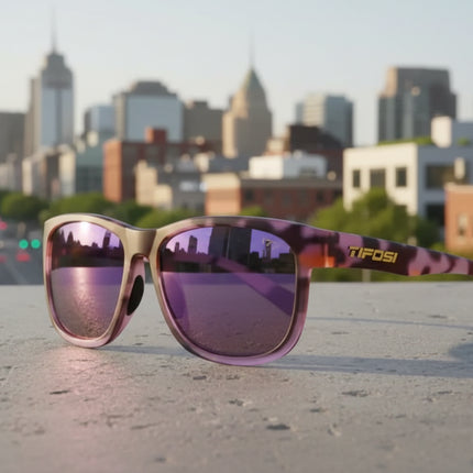 Tifosi sunglasses with purple lenses and a patterned frame, resting on a concrete surface with a city in the background