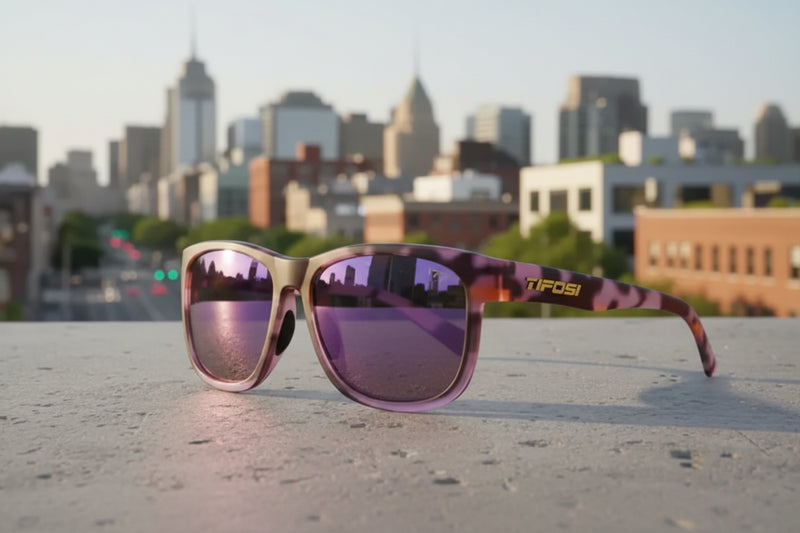 Tifosi sunglasses with purple lenses and a patterned frame, resting on a concrete surface with a city in the background