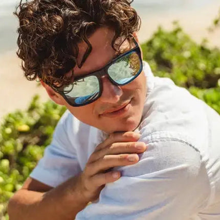 Man in sunglasses and white shirt on a beach with green foliage