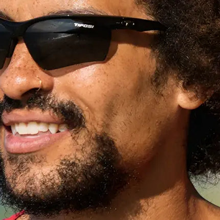 Man with curly hair and beard wearing black sunglasses with Tifosi logo