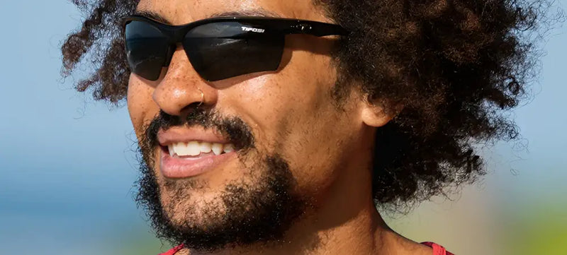Man with curly hair and beard wearing black sunglasses with Tifosi logo