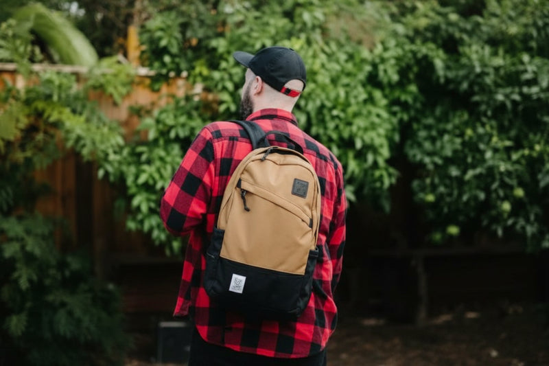 Man wearing a red and black plaid shirt and a black baseball cap, with a tan and black backpack