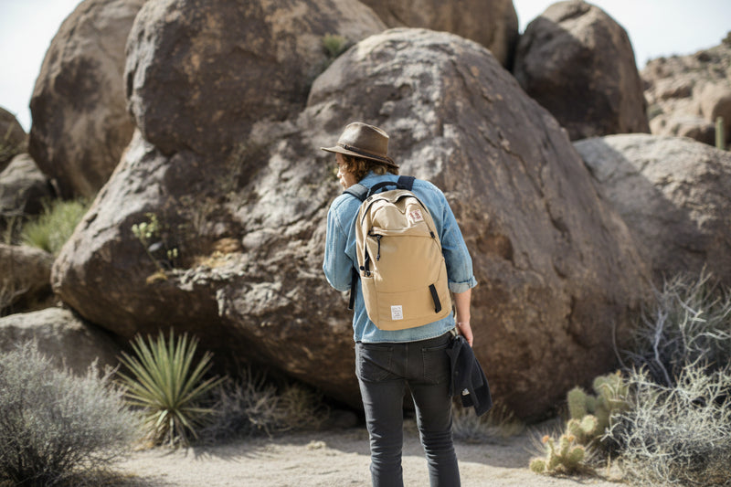 Man in a hat and denim shirt with a tan backpack, walking among large rocks and desert plants