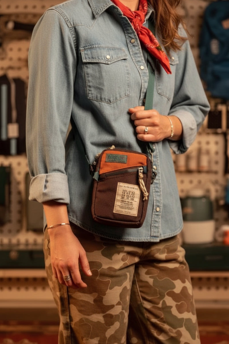 Person wearing a brown and orange crossbody bag over a light blue denim shirt and camouflage pants