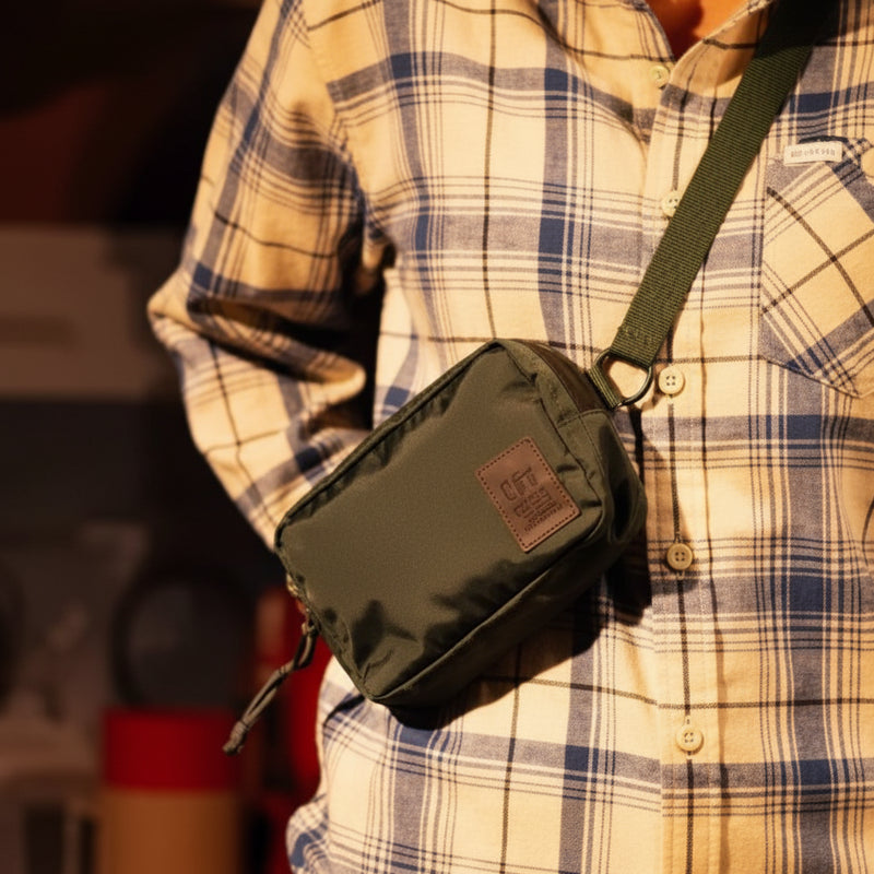 Person wearing a plaid shirt and a dark green crossbody bag with a brown leather patch
