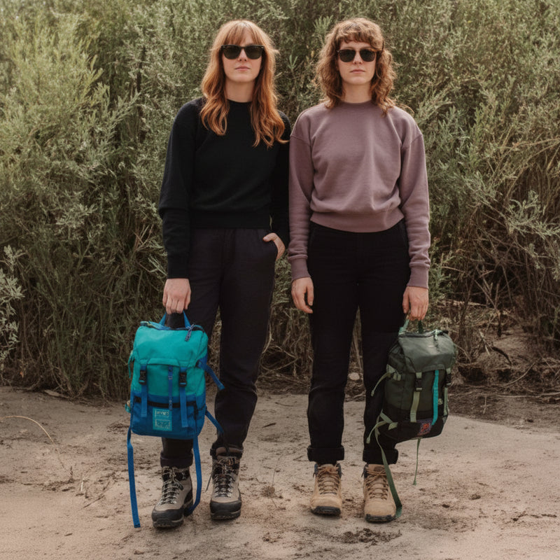 Two women in sunglasses and hiking gear stand with backpacks in a desert landscape