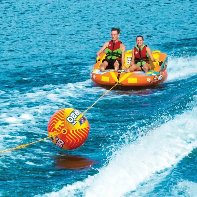 Two people in life vests on an orange inflatable tube being pulled by a rope with a yellow buoy